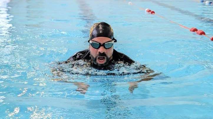 Kevin Duala is mid swim and has just taken his head out of the water for a breath of air. He has a black beard and is wearing a black swimming cap, goggles and a black shirt. His mouth is open wide as he take a breath before putting his head back underwater to continue his swim stroke. 