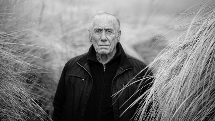 Black and white picture of Richard Lee. He is dressed in a black sweater and jacket between tufts of long grass in sand dune