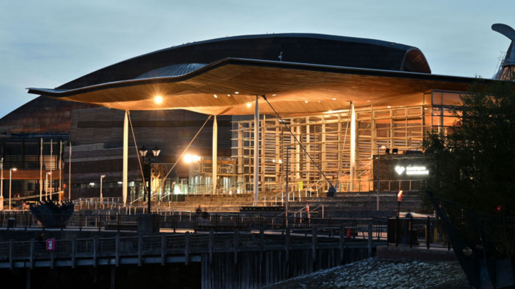 An exterior picture of the Senedd building with its lights on in the early evening.