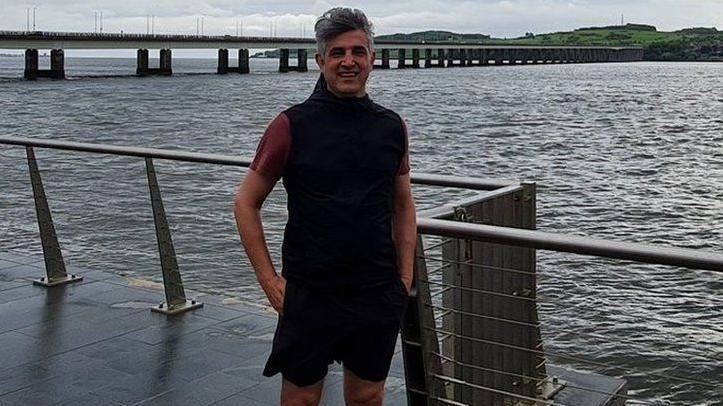 A man standing on a quayside in front of a river. In the background we can see a long car bridge crossing the river. He is wearing a black top and black shorts. It's an overcast day. 