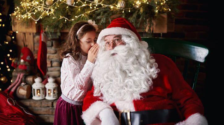 A young girl visits Santa and whispers into his ear. There are fairy lights and Christmas decorations behind them and red ribbons.