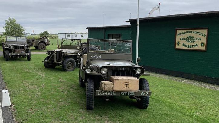 The outside of the 306th Bombardment Group Museum, a green box building, with a sign on the side of it, with three military vehicles by the side of it, on green grass. 