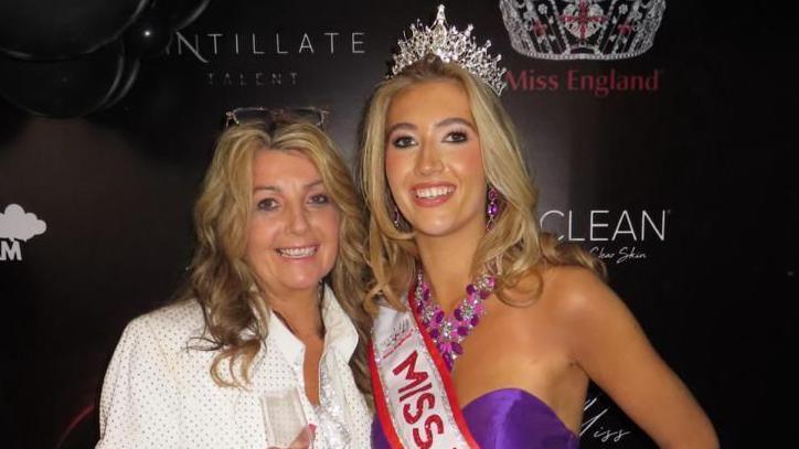 Tabitha and her mother Vicky stand in front of a black backdrop with small sponsorship logos on it. She wears a purple off the shoulder gown with large, puffy sleeves. She has long blonde hair and wears a silver crown and a red and silver sash. Vicky has long blonde hair and wears a white shirt.