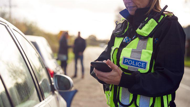 A woman police officer deals with a traffic incident and uses a mobile phone. Her uniform and the ends of her hair are visible. A car is parked on the verge and people are standing nearby.