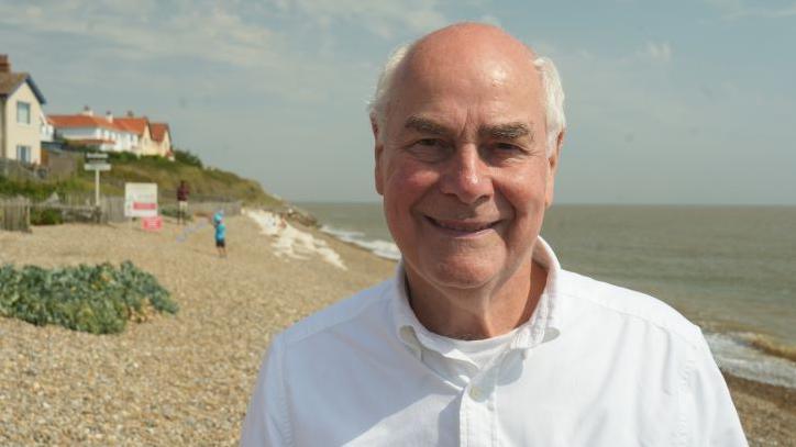 Mark Packnard is standing on the beach at Thorpeness in Suffolk. He is looking directly at the camera and is smiling. He is wearing a white shirt. The picture has been taken on a sunny day, there is a pebble beach and waves of the sea is crashing onto the beach.