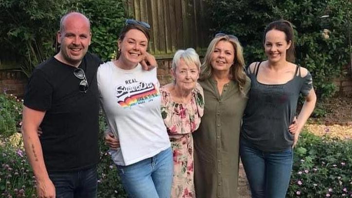 A man in a black t-shirt stands in a garden beside four women of varying ages. They all stand in a line with an arm over each other's shoulder in an embrace. They all smile for the photograph on a sunny day.