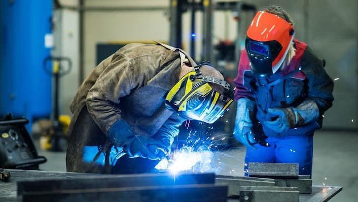 A man leans over a metal object that he is welding.  He has a full head visor on and the welding machine is creating white blue sparks.  A person stands leaning over him watching his work.