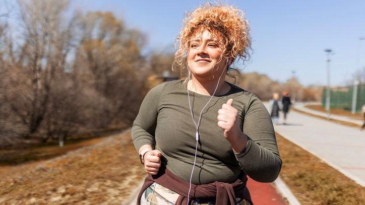 A woman with curly hair tied up in a ponytail is running outside in the sunshine, looking happy. She is wearing headphones and listening to music. People are walking nearby in the background.
