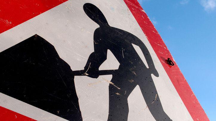 Red triangle sign with a black figure of a black shovelling on a white background with a blue sky behind it.