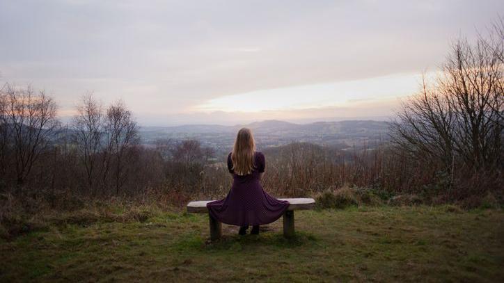 A woman with blonde hair and a polka dot dress sits on a bench with a view of the skyline across the Malvern Hills.