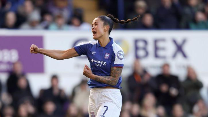 Natasha Thomas is pictured in the midst of a football game. She is celebrating with her right arm punching out. She is shout while jumping in the air. Supporters can be seen in the background watching on.