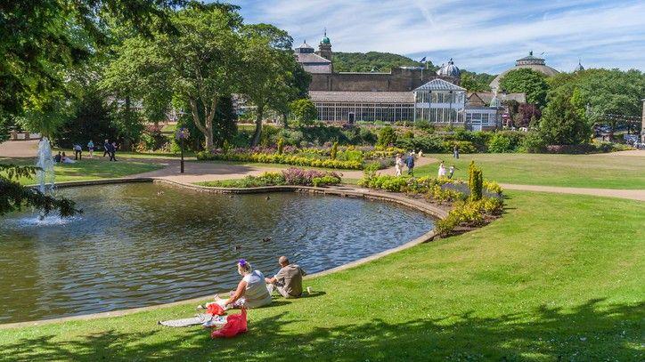 A couple sits on a lawn next to lake in the middle of a large park on a sunny day. Behind them is a large pavilion, orangery and dome-roofed building.