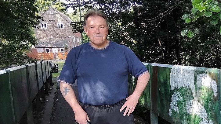 Councillor Phil Millward stands at one of the bridges over Hurley Brook. He is wearing a blue T-shirt and it is sunny. There are trees and a house in the background.