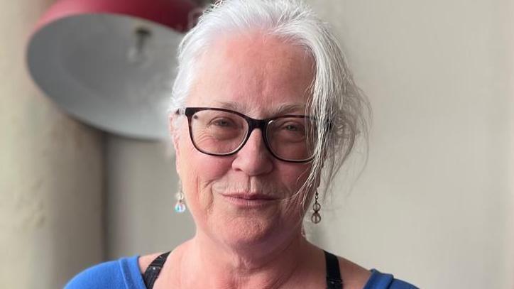 Majella Berrnad is wearing black-rimmed glasses and a blue top.
She has long hair and is wearing earrings.
She is standing in front of a large red lamp.