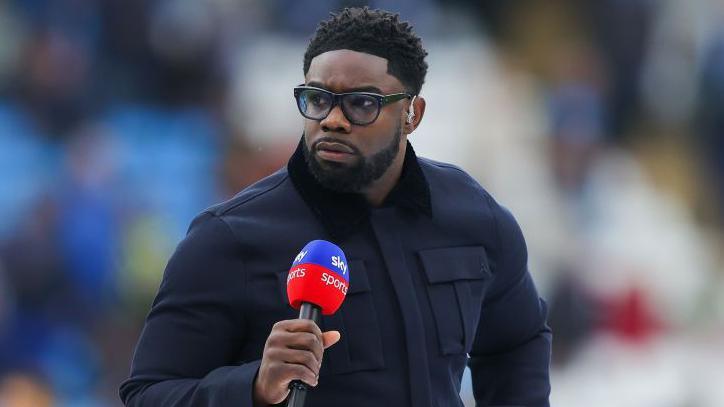 Micah Richards presenting for Sky Sports, he has short black hair and is wearing a utility style navy coat and black glasses