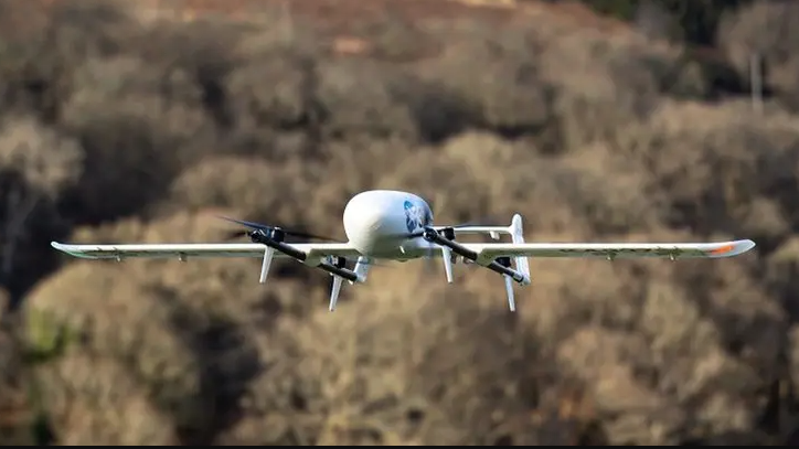 A white drone with wings and propellers flying in front of a wooded area.