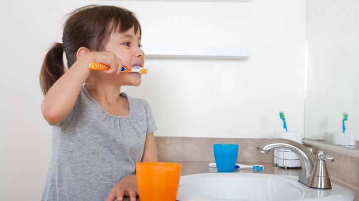 A girl brushes her teeth in front of a sink. There are blue and orange plastic beakers on the surface and there is another toothbrush in a holder by the taps. She is looking into the mirror as she cleans her teeth.