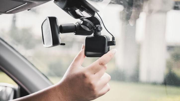 Video recording equipment in a car that has been set up on the rear-view mirror. The photograph is a close-up of the driver's hand adjusting the kit. The view outside the car is blurred.