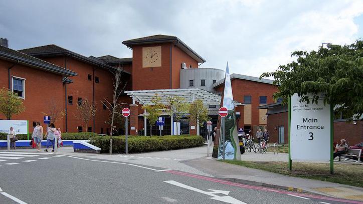 The main entrance to Wythenshawe Hospital. A road can be seen leading past the entrance. A sign can be seen with people walking over a crossing, some gathered by a bicycle racks and one sat on a bench. The red brick hospital building looms above the scene with a clock seen on the central tower.