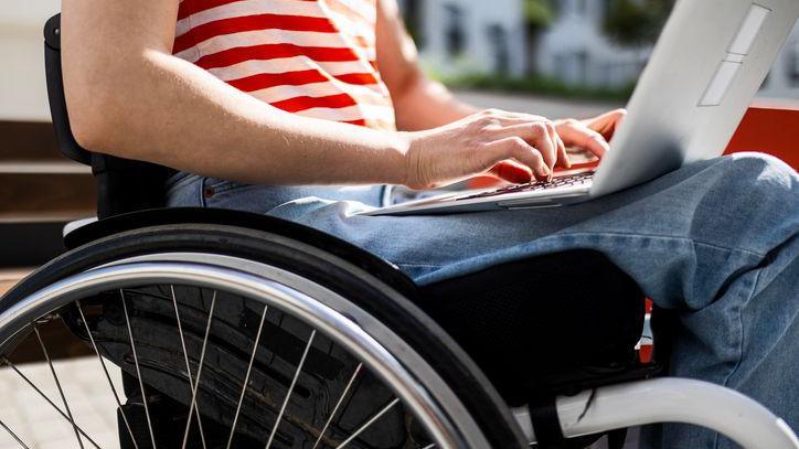 A close-up image of a wheelchair as a woman sits in and works on a laptop. Her jeans and red and white T-shirt and hands and forearms are visible.
