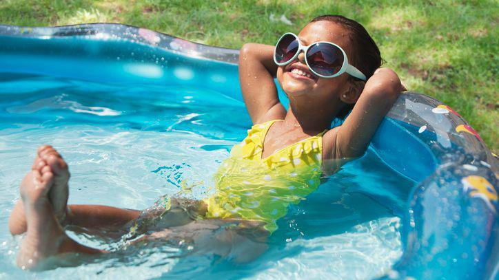 A girl wearing sunglasses smiling and relaxing in a paddling pool on a sunny day.