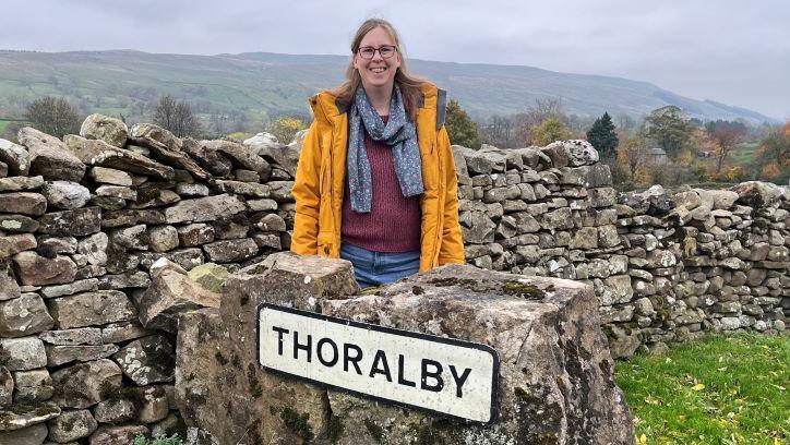 A woman in a mustard coat with a grey speckled scarf and maroon jumper stands next to a dry stone wall behind a village sign which says "Thoralby".