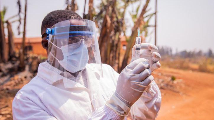 A medic in full protective face mask and white clothing, standing outside, holding up a syringe with gloved hands