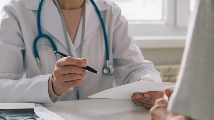 A woman GP has a consultation with a patient. She is sitting at a desk in front of a computer screen and has a stethoscope around her neck and is holding a pen and paper.