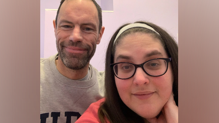 The image shows Alison Wright smiling directly at the camera. She is wearing glasses and has long, dark hair. Behind her, and also smiling at the camera, is her husband James. He has a dark beard and short hair.