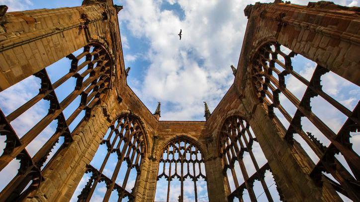 A bird flies over the bombed-out ruins of the Cathedral Church of St Michael in Coventry. The structure was destroyed in an air raid during World War Two. Walls, arches and decorative stone features still remain.