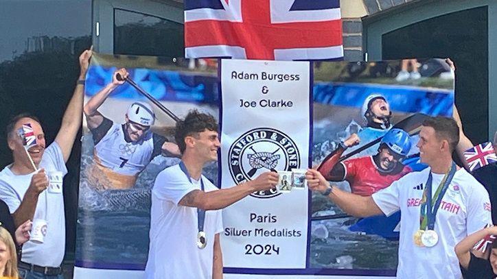 Two men wearing Olympic medals around their necks are clinking mugs together in "cheers". There is a banner behind them, with photos of them in their canoes. The banner reads "Adam Burgess and Joe Clarke. Paris silver medallists 2024." There is a great Britain flag in the background