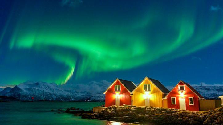Aurora Borealis over snowcapped mountains and fishermen cabins by the sea, Sommaroy, Troms og Finnmark, Norway