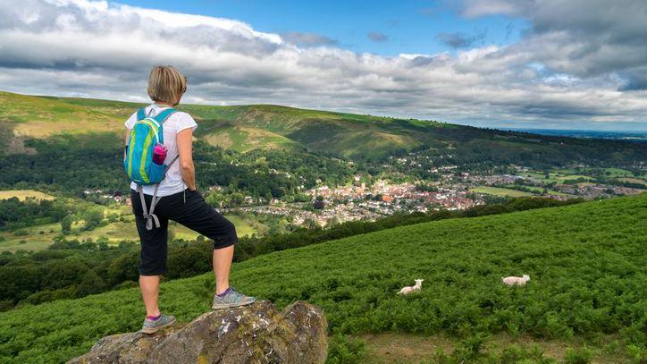 A woman hiking in the countryside looks at the view over Church Stretton and The Long Mynd from Ragleth Hill in the Shropshire Hills. She is standing on a rock and wearing a T-shirt and has a rucksack on her back. The town can be seen in the distance.