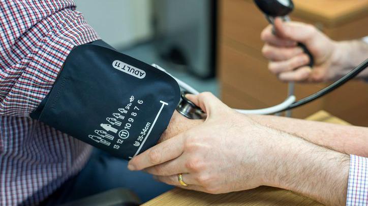 Stock close-up photo of an arm wearing a black blood pressure monitor. The arm has a red and white chequered shirt. The blood pressure is being taken by another arm which is holding down on the blood pressure monitor.