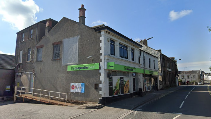The Co-op store in Aspatria is two storey building has a white facade with the lower ground displaying lime green signs for the shop and adverts on its windows. A rusting ramp at the side of the building leads to a windowless door. It is on the main street through Aspatria but there is very little activity.