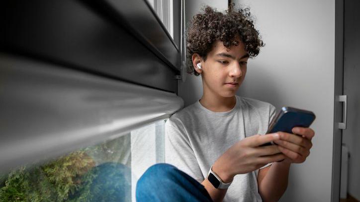 Teenage boy sitting at the window at home using mobile phone.