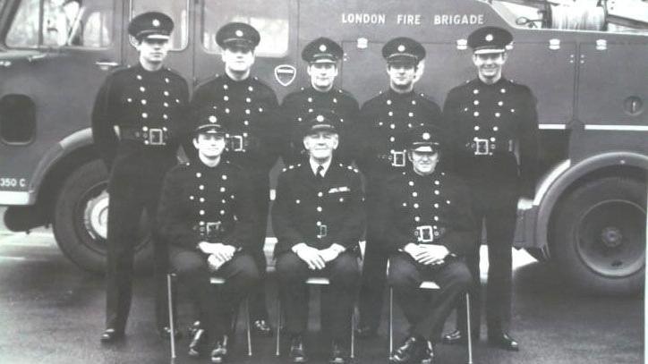 A black-and-white photograph of a group of eight male firefighters, stood in front of a London Fire Brigade fire truck. There are three firefighters sat on chairs in the front row, with five firefighters standing behind them in a row. They are all wearing matching outfits, with smart jackets, trousers and shoes, and a firefighters' cap. The truck behind them is positioned side-on.