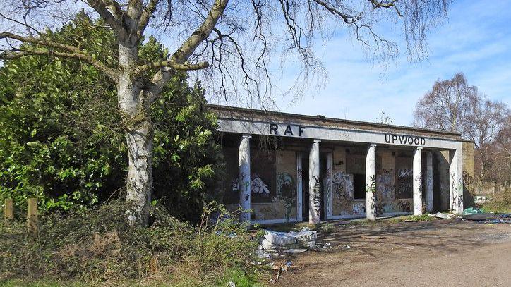 White single-storey building with "RAF Upwood" in black letters above the entrance. The facade has six pillars running from floor to roof. There is graffiti on the pillars. Wooden boards are visible behind the pillars. There are trees either side of the building.