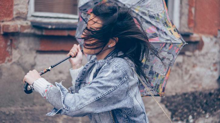 A woman holds an umbrella in the wind but it has blown inside out. Her hair is in her face and she is holding the umbrella with both hands in severe weather.