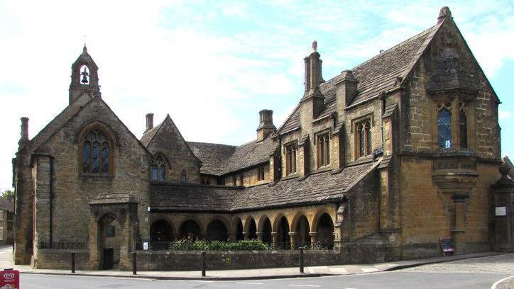 One side of St Johns' Almshouse on a street corner. The ornate, brown building includes a cloister, large arched windows and a rooftop bell.