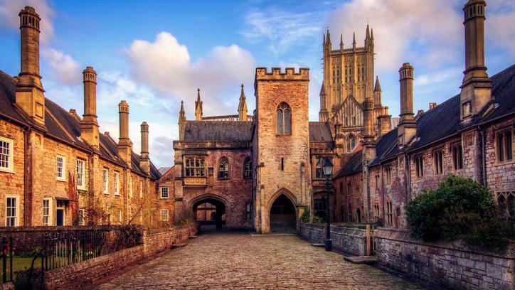 A cobbled street with terraces of medieval houses either side, a turreted gateway at the end and the cathedral tower visible in the background