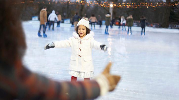 A girl skates to her mother smiling on an ice rink. She is wearing a white coat and white at. People are blurred in the background.