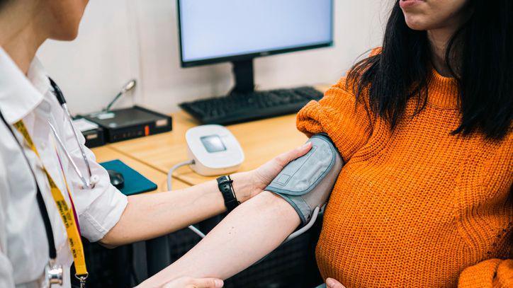 A GP, wearing a white shirt, takes the blood presure of a pregnant woman with dark, shoulder-length hair and who is wearing an orange jumper