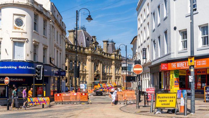 An urban renewal project takes place in Wolverhampton in 2022. A road is closed for construction work and people are walking around the city. There are shops on both sides of the road, and there is a sign saying "businesses open as usual".