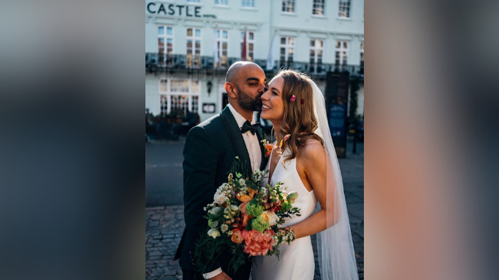 A bride and groom posing for a wedding picture in front of the The Castle Hotel in Windsor, which can be seen in the background. The groom is killing the bride on her right cheek. She is smiling, holding a bouquet of flowers.