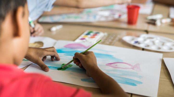A young child paints a picture. He is wearing a red top, and the picture is of the sea. 