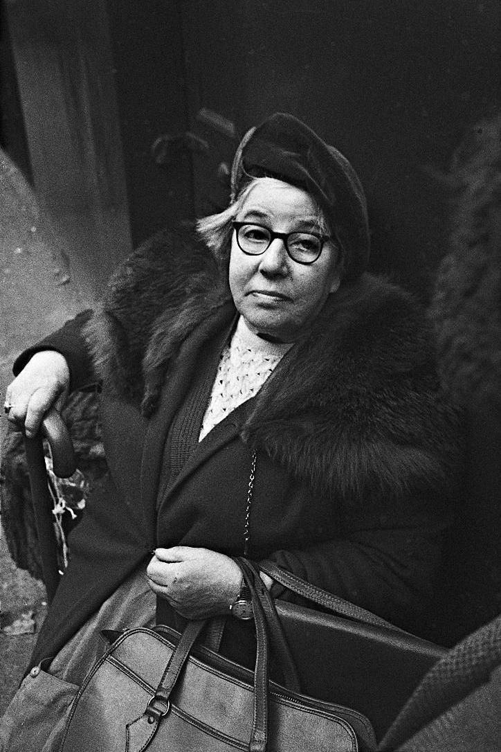 A black and white image of an elderly lady who is sitting down outside. She is looking into the camera and she is dressed in her best Sunday clothes with a dark coloured hat. 