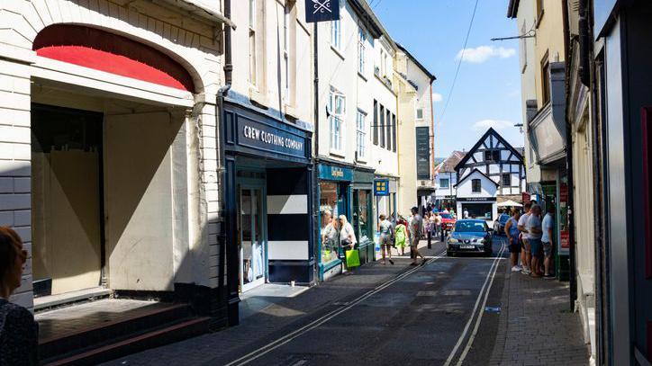 A street in Ludlow town centre. There are shops on either side of the narrow road, which has double yellow lines, and people walking on the pavements. The road leads to a Tudor-style black and white building.