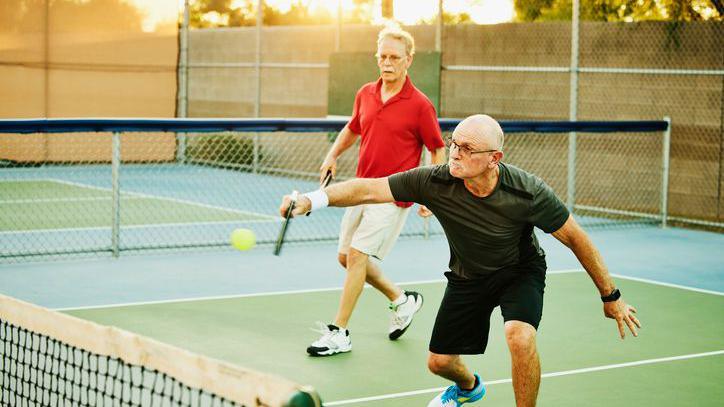 Wide shot of a man hitting backhand shot at net while playing doubles pickleball on summer evening 