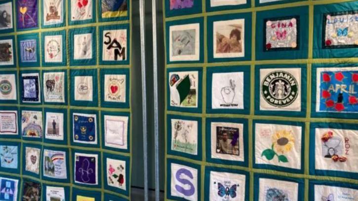 A shot of a memorial quilt in Hampshire made up of lots of individually decorated squares. It has a green background and individual square designs. 